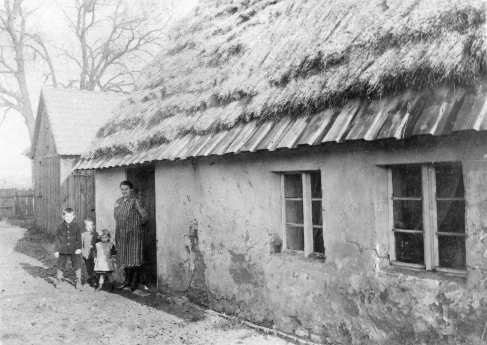 Rok i miejsce wykonania: jesień 1929 r., Chróścice, gm. Dobrzeń Wielki, pow. opolski, woj. opolskie. Fotografia przedstawia kobietę w codziennym odzieniu domowym, stojącą z trójką małych dzieci przy wejściu do domu. W tle widoczne zabudowania gospodarcze. Z załączonych informacji wynika, że zdjęcie wykonano przed domem rodzinnym Sopów. Autor zdjęcia: fotograf Karol Weis, właściciel atelier w Opolu z filą w Krapkowicach. Zdjęcie ze zbiorów prywatnych rodziny Sopa. Nr kat. 19-4