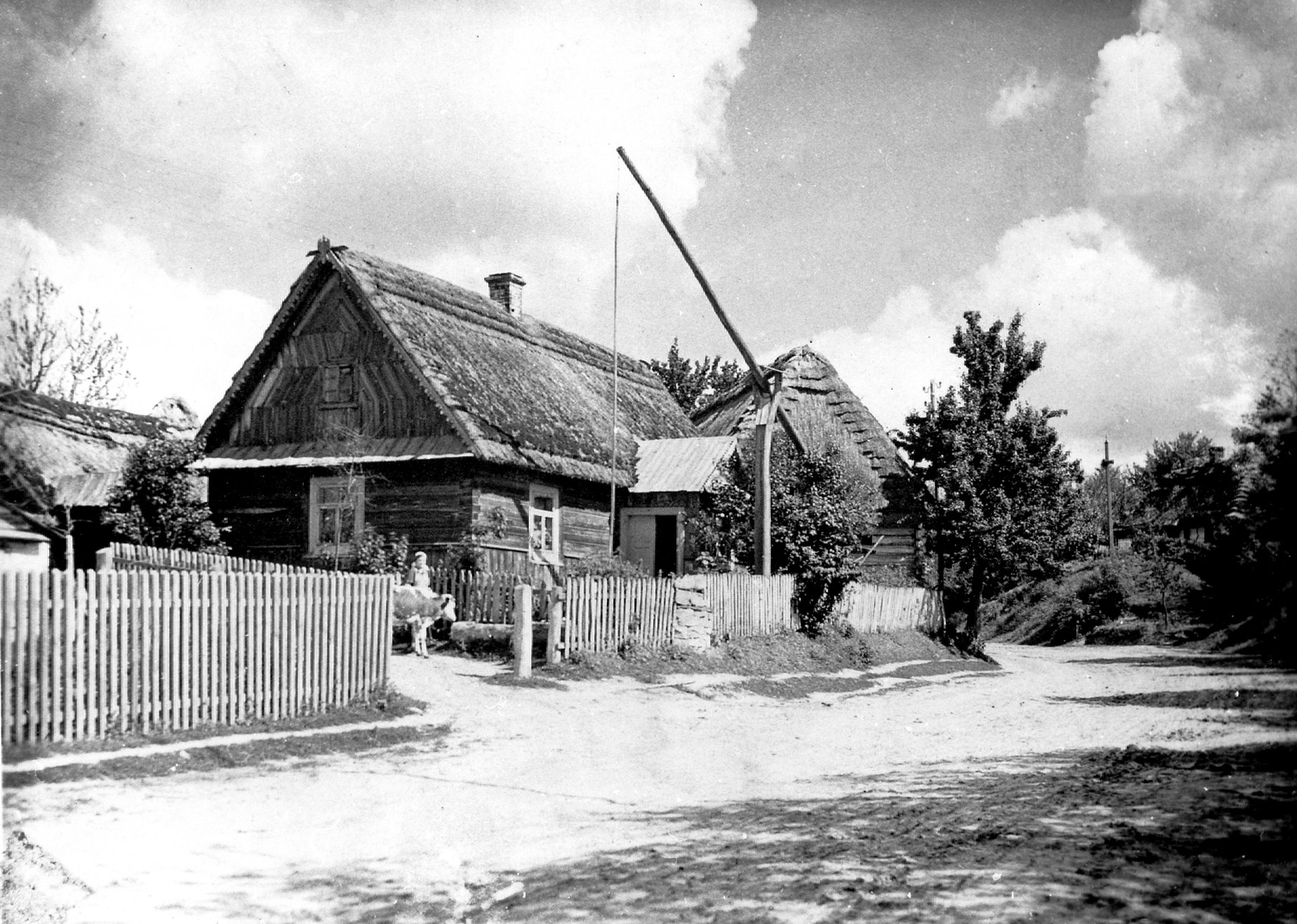 Rok i miejsce wykonania: maj 1940 r., wieś Zaburze, gm. Radecznica, pow. Zamość, woj. lubelskie. Fotografia przedstawia widok na siedlisko rodziny Kruczkowskich z Radecznicy. Zabudowania gospodarcze stoją przy drodze wiejskiej, w tle widać studnię z żurawiem, a w głębi przydrożny krzyż oraz koryto do pojenia zwierząt, zrobione z wydrążonego pnia drzewa. Autor zdjęcia: Pan Szymański (imię nieznane), fotograf-amator, który był wysiedlony z Wągrowca i przebywał okresowo w Radecznicy, pow. Zamość. Z zawodu był technikiem budowlanym, a zdjęcia robił hobbystycznie. Pod koniec maja 1940 r. został z żona i córeczką aresztowany przez Gestapo i wszyscy zginęli w obozie. Zdjęcie ze zbiorów prywatnych, nadesłał: Jan Kruczkowski z Zaburza, pow. Zamość, woj. lubelskie. Nr kat. 57-1