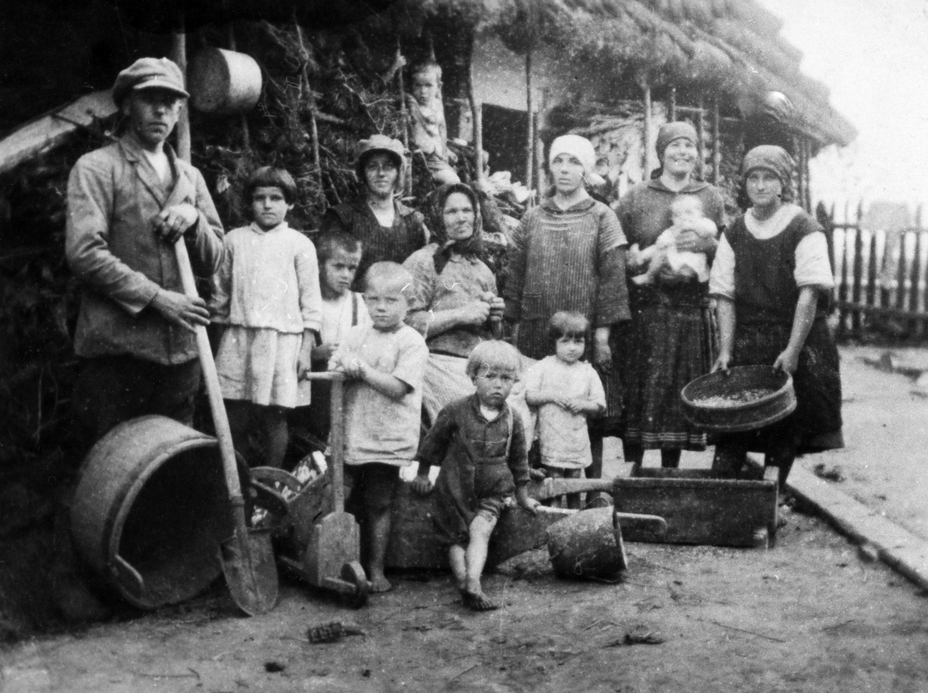 Rok i miejsce wykonania: lato 1929 r., Białobrzegi, gm. Białobrzegi, pow. łańcucki, woj. podkarpackie.

Fotografia przedstawia pięć kobiet w różnym wieku, mężczyznę i siedmioro dzieci, w tym niemowlę. Grupa stoi na podwórzu przed wiejskim domem krytym strzechą. Na ziemi przed ludźmi poukładane są różne gospodarskie narzędzia oraz naczynia. Przed mężczyzną leży dzieża, a on wspiera się na stylu od łopaty. Obok niego stoi dziewczynka w jasnej bluzce i spódniczce oraz chłopiec, który trzyma drewnianą dziecięcą zabawkę. Widoczny jest tylko jej przód, ale przypomina hulajnogę. W środku grupy siedzi najstarsza kobieta, trzymająca w dłoniach trudny do rozpoznania przedmiot. Wszyscy ubrani są w skromną, codzienną odzież. Jedna z kobiet ma na głowie kaszkiet, pozostałe noszą chustki zawiązane z tyłu głów, z wyjątkiem najstarszej, która zawiązała ją pod brodą. Dzieci są bez obuwia. Niemowlę w białej koszulce trzyma na rękach uśmiechnięta kobieta. Jedno z młodszych dzieci wspięło się na drewno ułożone wzdłuż ścian domu (zapewne zgromadzone na opał).

Według relacji nadawczyni zdjęcia przedstawia ono: „spotkanie w porze obiadowej na podwórzu u sąsiadów Wojnarów. Gospodynią tam była znana i lubiana w okolicy kobieta (na zdjęciu najstarsza kobieta obierająca kartofle). Obok niej w kaszkiecie stoi jej córka Kasia – krawcowa, sito trzyma druga córka Zośka. Jedyny mężczyzna, widoczny z lewej strony kadru, to kolejny sąsiad, a jego żona trzyma na rękach najmłodszą córeczkę Zosię. Młoda dziewczyna z sitem w ręce wyjechała na roboty do Francji i do kraju już nie wróciła. Dzieci żyją wszystkie oprócz małego blondynka na samym przodzie. Utopił się w czasie kąpieli w Wisłoku w czasie okupacji niemieckiej” (pisownia oryginalna). Autor zdjęcia: Franciszek Kulpa, ojciec nadawczyni. Dzięki niej dowiadujemy się więcej o Franciszku Kulpie. Był on fotografem chłopskim okresu międzywojennego. Pracował jako organista kościelny, a także kierował lokalną orkiestrą dętą. Z czasem jego prawdziwą pasją stała się fotografia. Wiele zdjęć zrobił starym aparatem na drewnianym statywie, niekiedy używał samowyzwalacza. Szczególnie lubił robić zdjęcia okazjonalne dzieciom z okolicznych wsi.

Zdjęcie ze zbiorów prywatnych, nadesłała: Danuta Drumlewska z Opola, woj. opolskie.

Nr kat. 250-2