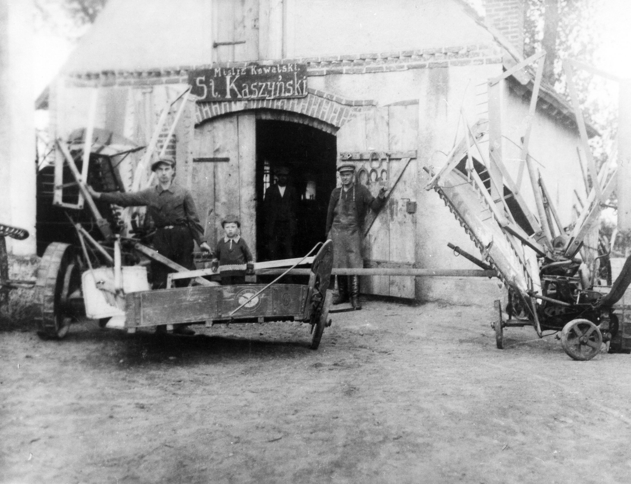 Rok i miejsce wykonania: 1944 r., Wylatowo, gm. Mogilno, pow. mogileński, woj. kujawsko-pomorskie.

Fotografia przedstawia podwórko przed warsztatem kowalskim, z jego pracownikami oraz małym chłopcem i różnymi narzędziami. Przed uwidocznioną na zdjęciu maszyną (żniwiarką) stoi mężczyzna z kilkuletnim chłopcem. W drzwiach warsztatu stoi drugi mężczyzna w roboczym fartuchu. Za nim widać wiszące na wieszaku podkowy różnej wielkości. W tle, po prawej i lewej stronie, stoją rozmaite sprzęty gospodarcze o niejasnym przeznaczeniu (prawdopodobnie przywiezione do naprawy). Nad bramą do warsztatu znajduje się szyld z napisem: „Mistrz Kowalski St. Kaszyński”. Wewnątrz budynku, w zacienieniu, stoi jeszcze jeden, słabo widoczny mężczyzna.

Na odwrocie zdjęcia napis: „Warsztat męża, Stefana Kaszyńskiego z firmą. Przy żniwiarce zbóż uczeń z najmłodszym synkiem. Fotka z 1944 roku, Wylatowo” (pisownia oryginalna).

Autor zdjęcia: nieznany.

Zdjęcie ze zbiorów prywatnych, nadesłała: Maria Kaszyńska z Wylatowa, pow. mogileński, woj. kujawsko-pomorskie.

Nr kat. 285-35