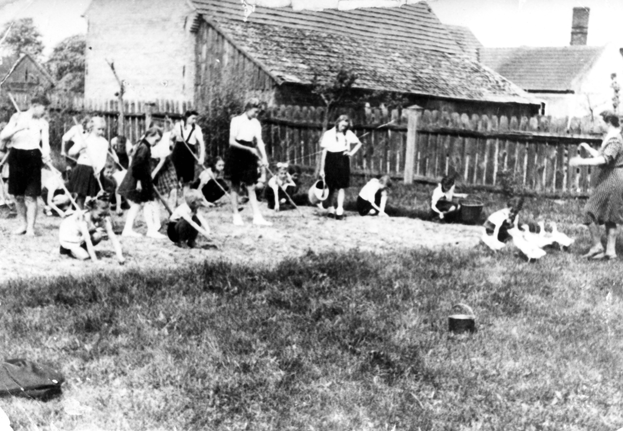 Rok i miejsce wykonania: 1946 r., Chrośnica, gm. Zbąszyń, pow. nowotomyski, woj. wielkopolskie.

Fotografia przedstawia grupę dziewcząt w białych bluzkach i ciemnych spódniczkach, pracujące na poletku. Niektóre grabią, inne pielą grządki, a jedna z nich z konewką podlewa rośliny. Po prawej stronie zdjęcia widać kobietę zaganiającą gęsi. W tle widoczne zabudowania gospodarcze i drewniany płot.

Według nadawczyni zdjęcia przedstawia ono wiosenne prace w szkolnym ogródku w Chrośnicy. Wśród uwiecznionych osób znajduje się mama właścicielki zdjęcia – drobna blondynka z grabiami, druga od lewej strony zdjęcia. Kobieta pędząca gęsi to wychowawczyni, Anna Siwińska.

Autor zdjęcia: nieznany.

Zdjęcie ze zbiorów prywatnych, nadesłała: Radosława Besińska z Chrośnicy, pow. nowotomyski, woj. wielkopolskie.

Nr kat. 148-9