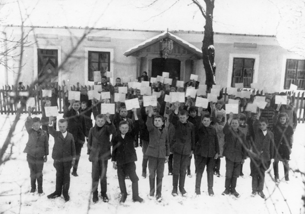 Rok i miejsce wykonania: zima, lata 30. XX wieku, Prandocin, gm. Słomniki, pow. krakowski, woj. małopolskie.

Fotografia przedstawia kilkudziesięcioosobową grupę chłopców, trzymających w rękach wyciągnięte do góry kartki. W tle widać podłużny, parterowy budynek z gankiem. Według nadawczyni zdjęcia przedstawia ono grupę dzieci szkolnych zgromadzonych przed starą szkołą w Prandocinie, w powiecie Miechowskim, prezentujących legitymacje przynależności do Kasy Stefczyka..

Autor zdjęcia: nieznany.

Zdjęcie ze zbiorów prywatnych, nadesłała: Halina Danilczyk z Krakowa, woj. małopolskie.

Nr kat. 120-6