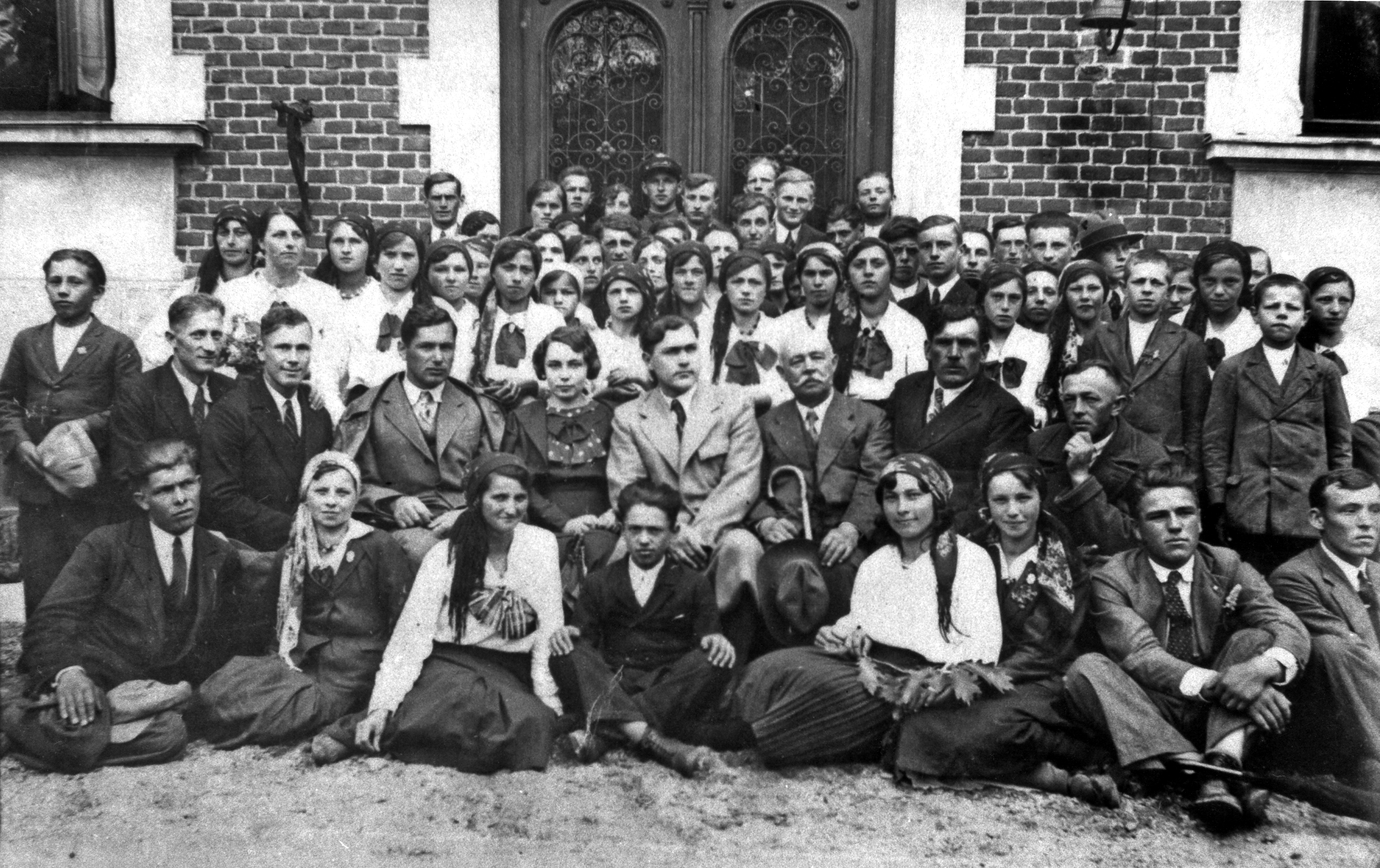 Rok i miejsce wykonania: 1935 r., Miłocin (obecnie część Rzeszowa), pow. rzeszowski, woj. podkarpackie.

Fotografia przedstawia grupę młodzieży zgromadzoną przed okazałym, murowanym budynkiem; widoczne są ozdobne drzwi, a po ich bokach – fragmenty dwóch okien. W drugim rzędzie siedzi osiem najstarszych osób, w tym jedna dwudziestokilkuletnia kobieta. Jest ona elegancko ubrana i wyróżnia się na tle pozostałych (między innymi brakiem chusteczki na głowie). Wszystkie pozostałe kobiety i dziewczęta są elegancko ubrane w jasne bluzki i ciemne spódnice, a na głowach mają zawiązane z tyłu chustki. Męska młodzież jest ubrana w garnitury i białe koszule; część z nich ma krawaty. Dwie osoby wyróżniają się nakryciem głowy. Mężczyzna stojący w ostatnim rzędzie, pośrodku, ma na głowie rogatywkę (wojskową lub harcerską). Druga osoba, kobieta z prawej strony zdjęcia, ma na głowie nakrycie przypominające kapelusz skautowski.

Według właścicielki zdjęcia fotografia przedstawia Stowarzyszenie Młodzieży z Handzlówki na wycieczce do Szkoły Rolniczej w Miłocinie k. Rzeszowa w 1935 r. Kobieta stojąca w pierwszym rzędzie, trzecia od prawej, to mama właścicielki zdjęcia – Julia z Wojnarów Magrysiowa.

Autor zdjęcia: nieznany.

Zdjęcie ze zbiorów prywatnych, nadesłała: Maria Magryś z Rzeszowa, pow. rzeszowski, woj. podkarpackie.

Nr kat. 336-18