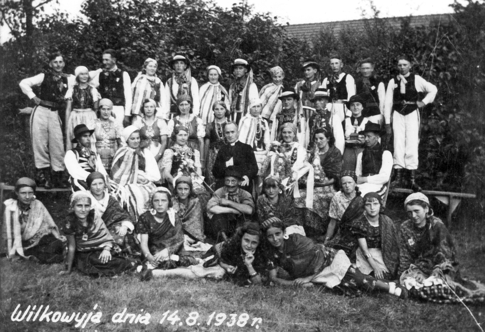 Rok i miejsce wykonania: 14.08.1938 r., Wilkowyja, gm. Jarocin, pow. jarociński, woj. wielkopolskie.

Fotografia przedstawia grupę młodzieży w bogatych strojach ludowych i siedzącego pośrodku nich księdza. Według nadawczyni zdjęcia, zostało ono wykonane z okazji dożynek i „święta plonów”. Przedstawia młodzieżowy zespół ludowy z Wilkowyi, w którym występowała również jej matka, Stanisława Bartkowiak. Zdjęcie wykonano na terenie plebanii, stąd też obecność księdza na honorowym miejscu.

U dołu zdjęcia napis: „Wilkowyja dnia 14.08.1938 r.”.

Autor zdjęcia: nieznany.

Zdjęcie ze zbiorów prywatnych, nadesłała: Grażyna Kaczmarek z Wilkowyi, pow. jarociński, woj. wielkopolskie.

Nr kat. 62-1