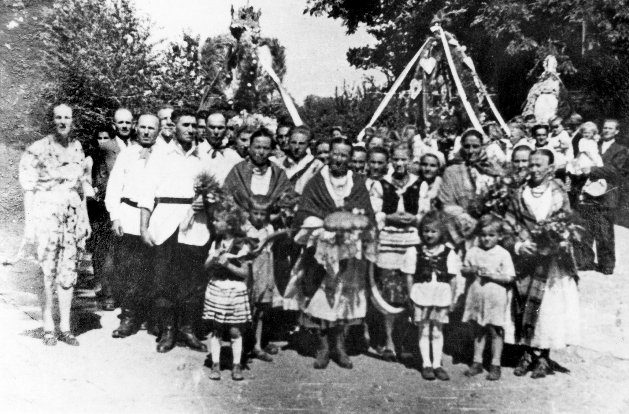 Rok i miejsce wykonania: 1936 r., Sobniów, gm. Jasło, pow. jasielski, woj. podkarpackie.

Fotografia jest grupowym ujęciem uczestników dożynek. Na pierwszym planie widoczne są osoby ubrane w ludowe stroje. Kobiety mają na sobie białe bluzki, ciemniejsze spódnice, białe fartuchy, a na szyjach korale. Niektóre mają również na głowach chustki oraz duże chusty zarzucone na ramiona. Dwie małe dziewczynki również ubrane są w strój ludowy, dodatkowo mają na sobie gorsety. Mężczyźni ubrani są w białe koszule wypuszczone na wierzch, na ciemne spodnie. Koszule mają przepasane ciemnymi pasami, a nogawki spodni wpuszczone w wysokie czarne buty. Na przodzie pochodu, w centralnym miejscu, stoi kobieta trzymająca w rękach bochen okrągłego chleba, ułożony na białej, ażurowej serwecie. Przed nią stoją dzieci, a obok inne kobiety i mężczyźni w strojach ludowych. Pozostali uczestnicy dożynek, stojący dalej, ubrani są w odświętne, miejskie ubrania. Za ludźmi i ponad nimi widoczne są trzy duże wieńce dożynkowe; jeden z nich ma kształt korony.

Autor zdjęcia: nieznany.

Zdjęcie ze zbiorów prywatnych, nadesłała: Beata Wójcik.

Nr kat. 324-1