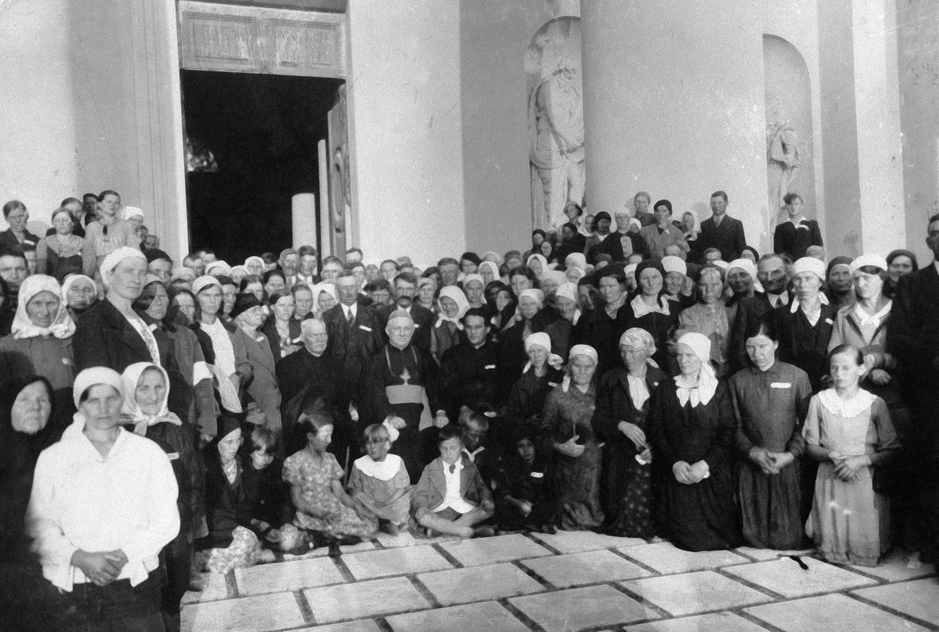 Rok i miejsce wykonania: 1937 r., Wilno, obecnie Litwa.

Fotografia przedstawia około stuosobową grupę ludzi pozujących do zdjęcia na dziedzińcu świątyni. W pierwszym rzędzie wiele osób klęczy. Pośrodku grupy siedzi trzech księży; wśród nich znajduje się biskup. U ich stóp na podłodze siedzi siedmioro dzieci. W grupie zdecydowanie przeważają kobiety. Prawie wszystkie mają głowy osłonięte chustkami. W tle widać rzeźby we wnękach i wielkie drzwi prowadzące do wnętrza świątyni. Według nadawcy zdjęcia przedstawia ono pielgrzymkę z parafii Boguty do Wilna w 1937 roku.

Autor zdjęcia: nieznany.

Zdjęcie ze zbiorów prywatnych, nadesłał: Roman Świerżewski z Drewnowa, pow. ostrowski, woj. mazowieckie.

Nr kat. 246-7