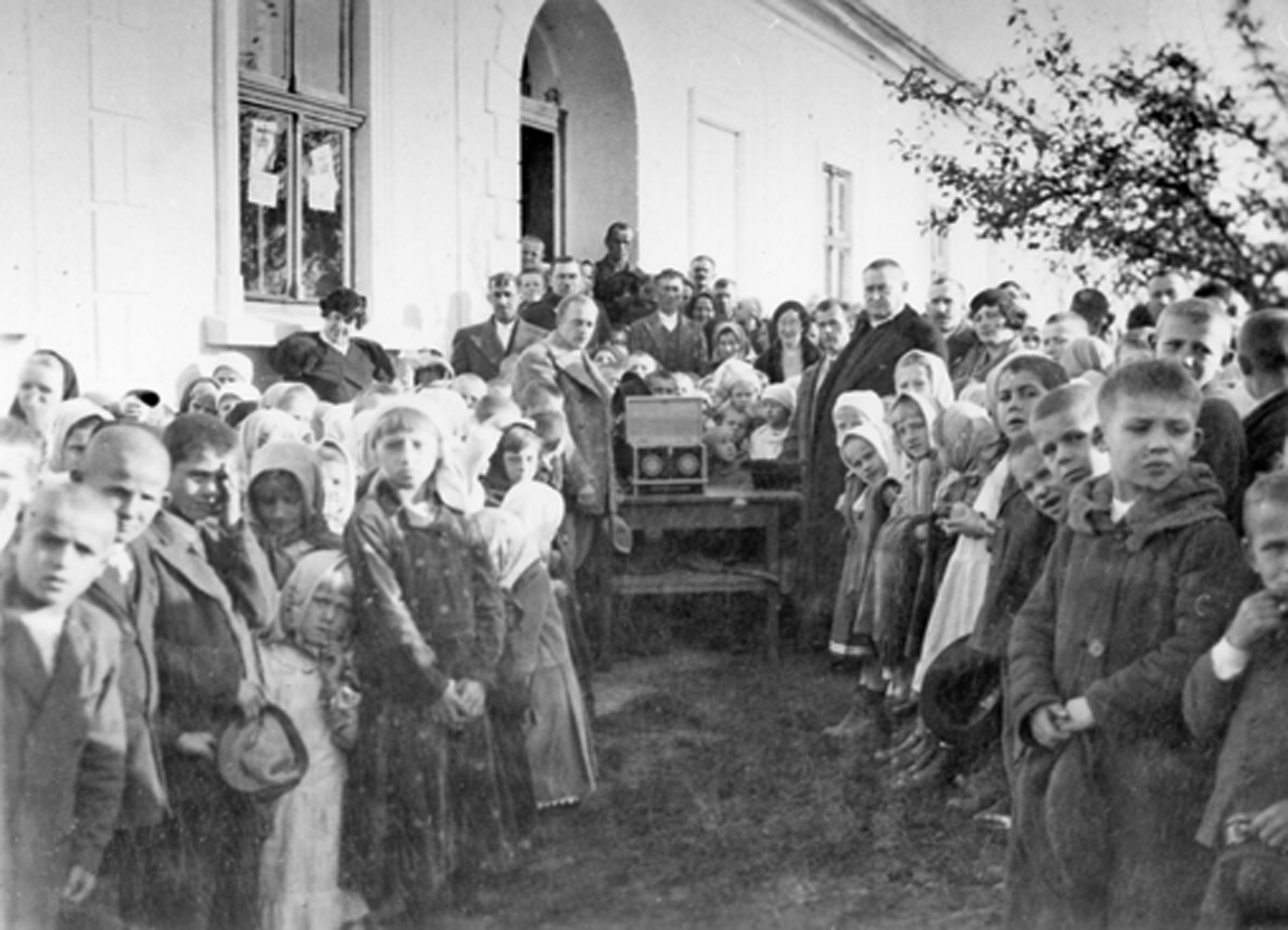 Rok i miejsce wykonania: 1935 r., Podgrodzie (właściwe: Podegrodzie), gm. Podegrodzie, pow. nowosądecki, woj. małopolskie.

Fotografia przedstawia liczną grupę osób, w tym wiele dzieci, stojących w dwóch rzędach przed wejściem do dużego murowanego budynku. Na pierwszym planie widoczne są przede wszystkim dzieci w różnym wieku, ubrane skromnie i raczej biednie. Dziewczynki mają na głowach jasne chustki, a chłopcy w rękach trzymają czapki. Wielu z nich ma ogolone głowy.

Według nadawcy zdjęcia dotyczy ono Podegrodzia i przedstawia pobranie ziemi na budowę kopca Marszałka J. Piłsudskiego w Krakowie. Ziemię pobrano z terenu plebani, gdzie w 1914 r. Marszałek kwaterował.

Autor zdjęcia: nieznany.

Na odwrocie zdjęcia napis: „Pobranie ziemi z pod plebanii w Podgrodziu, z miejsca gdzie w 1914 r. kwaterował marsz. Józef Piłsudski – na kopiec Marsz. J. Piłsudskiego w Krakowie. Podgrodzie 1935 r.” (pisownia oryginalna). Zdjęcie ze zbiorów prywatnych, nadesłał: Stanisław Chrząstowski z Podegrodzia, pow. nowosądecki, woj. małopolskie.

Nr kat. 193-24