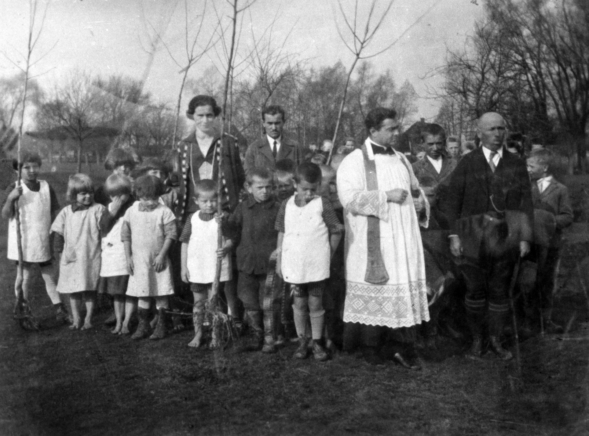 Rok i miejsce wykonania: 1927 r., Białobrzegi, gm. Białobrzegi, pow. łańcucki, woj. podkarpackie.

Fotografia przedstawia grupę kilkunastu dzieci wraz z dorosłymi: kobietą i czterema mężczyznami, z których jeden był księdzem. Dzieci widoczne na pierwszym planie odziane są w białe fartuszki nałożone na inne ubrania. Niektóre z nich nie mają butów. Kilkoro trzyma w rękach młode drzewka z widocznymi korzeniami, unosząc je nad ziemią. W tle widać zagajnik. Według nadawczyni zdjęcia przedstawia ono dzieci szkolne i nauczycieli podczas uroczystości święcenia drzewek, przeznaczonych do obsadzania drogi we wsi Białobrzegi. Osoby widoczne na zdjęciu to ksiądz Partykiewicz, kierownik szkoły Cieśla i nauczyciele: Kulpa oraz Samsonowicz.

Autor zdjęcia: Franciszek Kulpa, ojciec nadawczyni (więcej informacji o autorze zdjęcia zob. opis fotografii 250-2).

Zdjęcie ze zbiorów prywatnych, nadesłała: Danuta Drumlewska z Opola, woj. opolskie.

Nr kat. 250-7