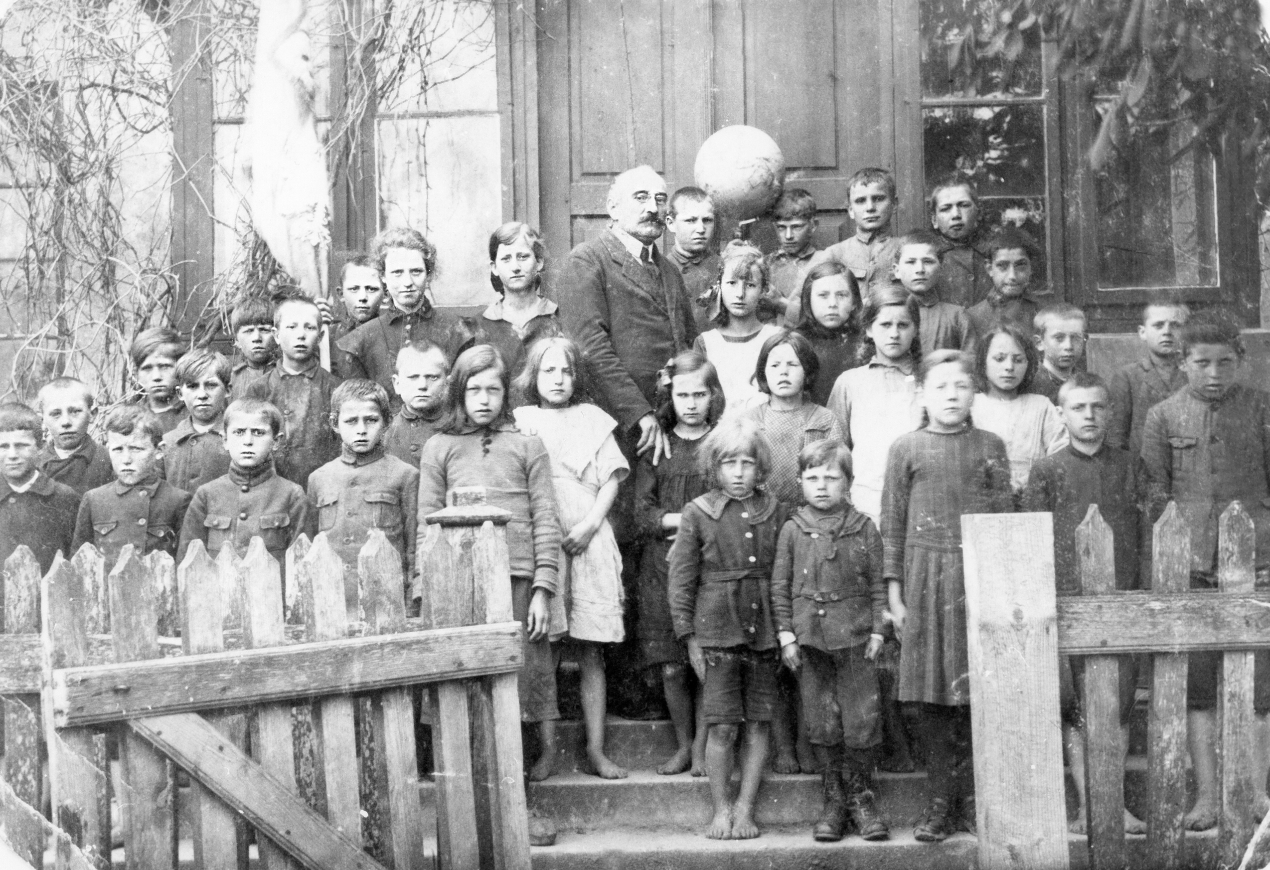 Rok i miejsce wykonania: 14.06.1923 r., Wieniec, gm. Brześć Kujawski, pow. włocławski, woj. kujawsko-pomorskie.

Fotografia przedstawia grupę dzieci w różnym wieku, stojących z nauczycielem przed budynkiem szkoły. Dzieci ubrane są biednie, wiele z nich nie ma butów. Część chłopców nosi bluzy mundurkowe; włosy mają ostrzyżone na krótko, a czasem nawet zgolone. Dziewczynki ubrane są w zwykłe codzienne sukienki. Włosy mają związane w warkocze lub (jeśli są na to za krótkie) rozpuszczone. Pomiędzy dziećmi stoi nauczyciel, a na lewo od niego młoda kobieta, również nauczycielka. W ostatnim rzędzie widoczny jest chłopiec, który trzyma globus. Z informacji na odwrocie zdjęcia wynika, że przedstawia ono uczniów klasy czwartej szkoły podstawowej we wsi Wieniec, w roku szkolnym 1922/1923. Wśród nich widoczny jest ojciec nadawcy, Józef Wesołowski, ur. 1911 r. i zm. 1966 r., stojący, jako „drugi na prawo od globusa”.

Rewers zdjęcia zadrukowany jest jak rewers pocztówki. W miejscu przeznaczonym na adres widnieje: „Jan Błasia Wieniec Włocławek”. W miejscu przeznaczonym na tekst znajduje się trudno czytelny napis: „Fot. Szkoły pow. (nieczytelne) IVtego w Wieńcu rok szkolny 14/VI 22/3 rok”.

Autor zdjęcia: Jan Besiak.

Zdjęcie ze zbiorów prywatnych, nadesłała: Kazimiera Michalska ze Szczecina, pow. Szczecin, woj. zachodniopomorskie.

Nr kat. 26-1