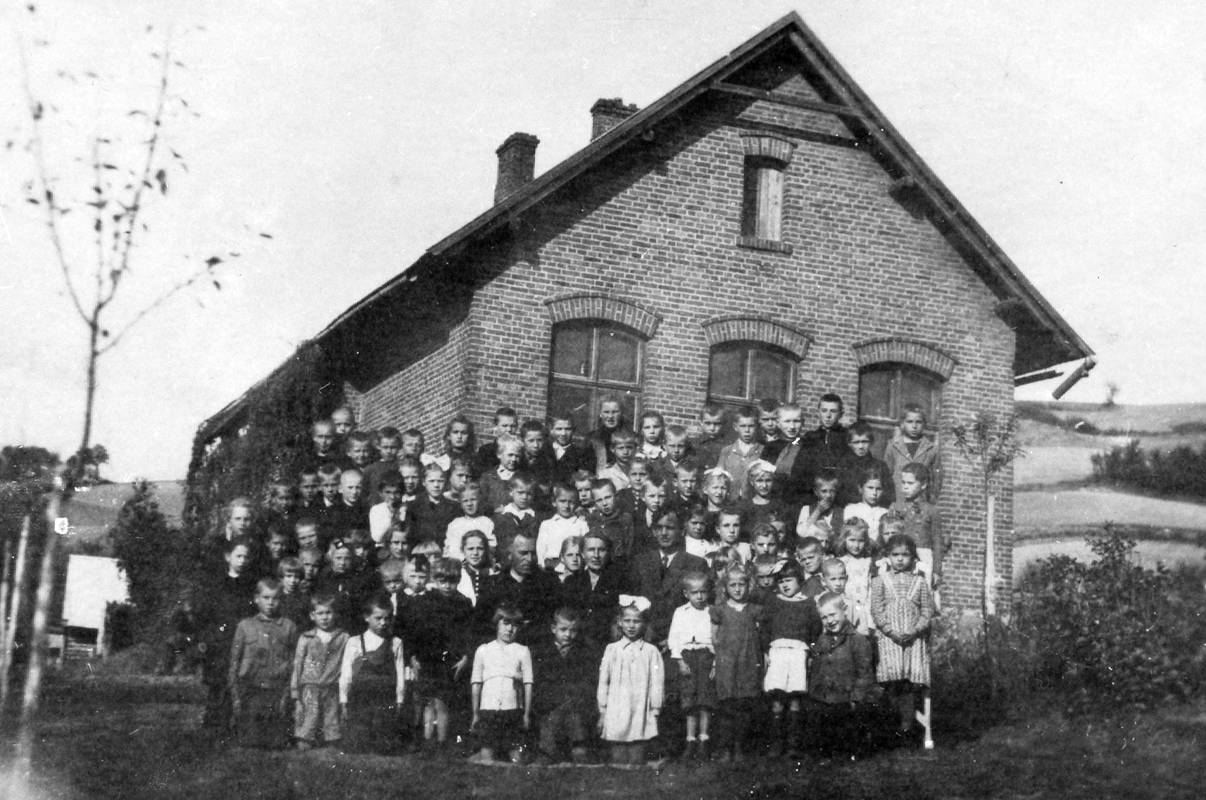 Rok i miejsce wykonania: 1945 r., Bukowa, gm. Brzostek, pow. dębicki, woj. podkarpackie.

Fotografia przedstawia grupę dzieci w rożnym wieku pozujących do zdjęcia przed dużym murowanym budynkiem szkolnym. Dzieci w pierwszym rzędzie klęczą. W drugim, pośrodku, siedzą nauczyciele: jedna kobieta i dwóch mężczyzn. Obok nich i za nimi stoją pozostałe dzieci, ubrane schludnie i starannie. Chłopcy często mają na sobie jasne koszule, ciemne marynarki lub kurtki, a dziewczynki sukienki bądź spodniczki. Włosy mają spięte, często przy tym związane kokardami. Nauczycielka ubrana jest w żakiet, a nauczyciele w garnitury i białe koszule z krawatami. Na odwrocie zdjęcia napis: „Stec Maria. Pamiątka z klasy trzeciej z Bukowej. Rok 1945”.

Autor zdjęcia: nieznany.

Zdjęcie ze zbiorów prywatnych rodziny Stec i Lechwar.

Nr kat. 60-6