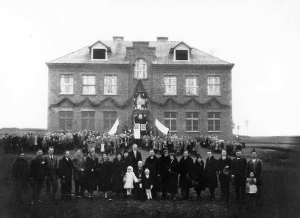 Rok i miejsce wykonania: listopad 1936 r., Prandocin, gm. Słomniki, pow. krakowski, woj. małopolskie.

Fotografia przedstawia duży, dwukondygnacyjny, murowany budynek z mansardami, zbudowany pośrodku łąki (pola). Przed budynkiem są licznie zgromadzeni ludzie. Część z nich –zapewne uczniowie – ustawiona jest w czterech grupach. Pośrodku, pomiędzy tymi grupami, pozostawiono szerszą, wolną przestrzeń, prowadzącą do głównego wejścia do szkoły. Na pierwszym planie, przed dziećmi szkolnymi, w jednym rzędzie stoi grupa dorosłych i troje dzieci.

Według informacji podanej przez właścicielkę zdjęcia są to członkowie Komitetu Założycielskiego. Wejście do szkoły jest przybrane roślinnymi girlandami, obrazami. Przestrzeń pomiędzy parterem i piętrem także jest przyozdobiona girlandami. Budynek nie jest jeszcze w pełni wykończony. Mansardy nie mają wstawionych okien, a szyby okien na piętrze wyglądają, jakby były czymś zasłonięte. Według właścicielki zdjęcia przedstawia ono uroczystość otwarcia Szkoły Powszechnej w Prandocinie. Pośrodku grupy Komitetu Założycielskiego i grona pedagogicznego, za siedzącym księdzem proboszczem kanonikiem Romualdem Wiadrowskim, nieco na prawo, stoją kierownik szkoły Marian Świebodziński oraz Ignacy Blitek, wójt-komisarz (po lewej stronie za księdzem). W skład Komitetu Założycielskiego wchodzili miejscowi chłopi, woj. Piotr Lupa, Kubuśka, Antoni Warchoł, Odrzywołek, Podsiadło, Jan Lech (sołtys), Michał Kopeć.

Autor zdjęcia: nieznany.

Zdjęcie ze zbiorów prywatnych, nadesłała: Halina Danilczyk z Krakowa.

Nr kat. 120-3