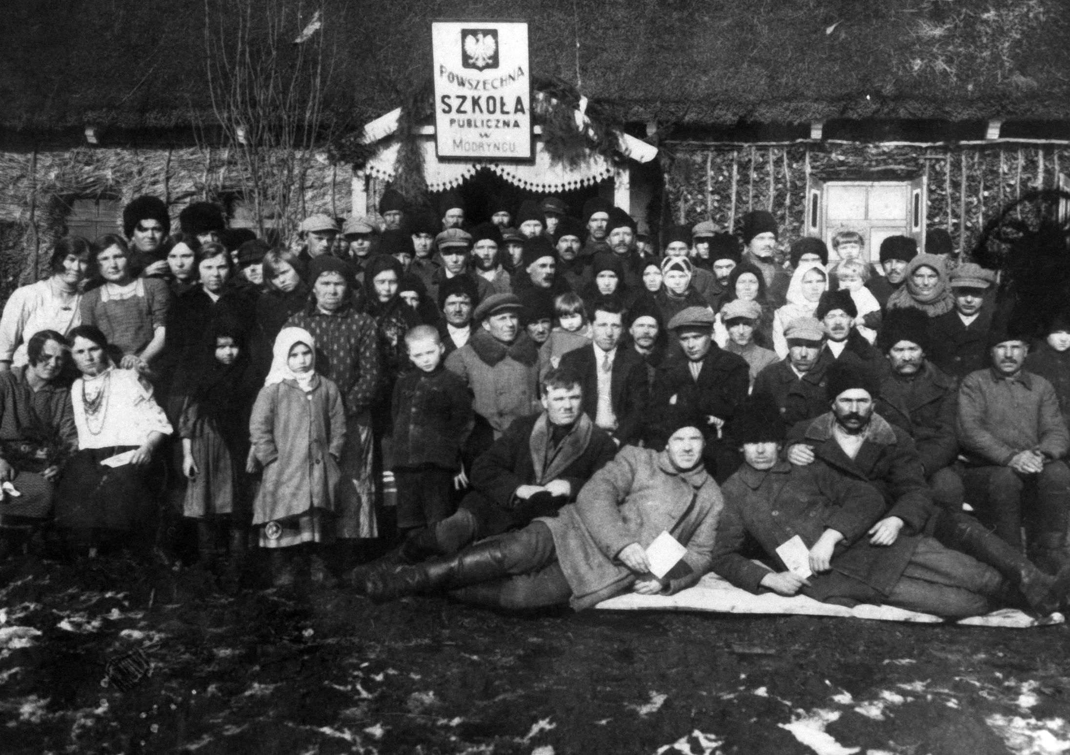 Rok i miejsce wykonania: 1930 r., Modryniec, gm. Mircze, pow. hrubieszowski, woj. lubelskie.

Fotografia przedstawia społeczność wiejską zebraną przed drewnianym budynkiem szkoły, o czym świadczy tablica przytwierdzona do ganku. Umieszczony na niej napis pod godłem brzmi: „Powszechna Szkoła Publiczna w Modryńcu”. Wśród zebranych ludzi widać również wiele dzieci. Wszyscy pozujący ubrani są w skromną, codzienną odzież wierzchnią: mężczyźni w grube kurtki-płaszcze i czapki, a kobiety w płaszcze i grube chusty. Cztery młode kobiety po lewej stronie zdjęcia pozują w samych sukienkach. Mężczyźni, którzy leżą w pierwszym rzędzie, trzymają w dłoniach książeczki lub kartki. Za nimi siedzi mężczyzna w garniturze, białej koszuli i krawacie. Zdjęcie wykonano wczesną wiosną. Na pierwszym planie widoczne są leżące na ziemi łachy śniegu.

Autor zdjęcia: nieznany.

Zdjęcie ze zbiorów prywatnych, nadesłał/nadesłała: brak danych.

Nr kat. 265-1