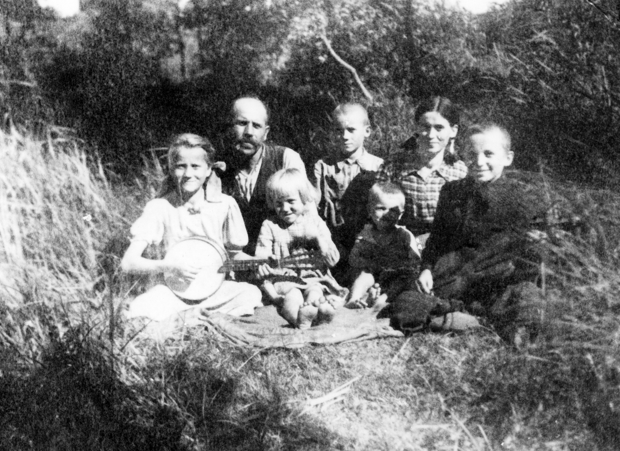 Rok i miejsce wykonania: 1942 r., Bychawa, gm. Bychawa, pow. Lublin, woj. lubelskie.

Fotografia przedstawia rodzinę z pięciorgiem dzieci siedzącą na kocu na łące. Dziewczynka, najstarsza z rodzeństwa, gra na instrumencie przypominającym mandolinę, a chłopiec siedzący po drugiej stronie koca trzyma w rękach skrzypce. Pomiędzy nimi siedzi dwójka najmłodszych dzieci, a za nimi – rodzice ze starszym synem. Wszyscy ubrani są schludnie i odświętnie. Dzieci i rodzice uśmiechają się, wyglądają na szczęśliwych. Dzień jest letni, słoneczny, a w tle widoczna bujna zieleń.

Z informacji nadesłanych przez nadawczynię zdjęcia wynika, że przedstawia ono jej babcię i dziadka oraz ich dzieci. Jest to rodzina ze strony jej ojca, która była bardzo muzykalna. Wszyscy trzej chłopcy grali na instrumentach muzycznych.

Autor zdjęcia: Zdzisław Nowakowski, mąż siostry babci.

Zdjęcie ze zbiorów prywatnych rodziny Kawała, Bychawa, pow. Lublin, woj. lubelskie.

Nr kat. 261-5