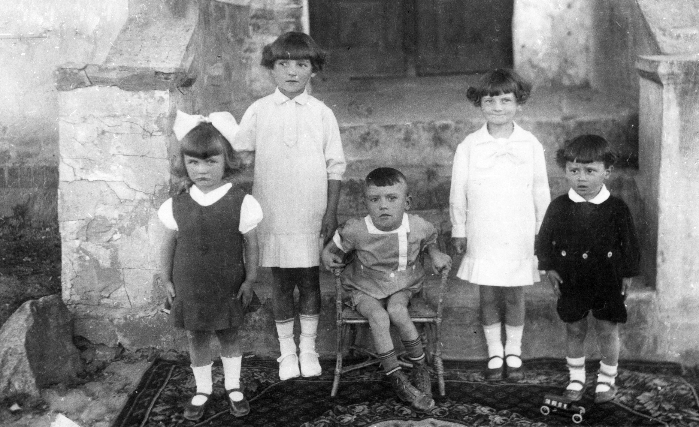 Rok i miejsce wykonania: 1936 r., Wylatowo, gm. Mogilno, pow. mogileński, woj. kujawsko-pomorskie.

Fotografia przedstawia pięcioro dzieci w różnym wieku pozujących do zdjęcia przed schodami prowadzącymi do budynku. Przed nimi, u ich stóp, leży wzorzysty dywan, na którym widać duży i efektowny, zabawkowy samochód. Dzieci ubrane są w odświętną odzież i stoją w szeregu wyprostowane do zdjęcia. Pośrodku, pomiędzy nimi, na wiklinowym krzesełku siedzi chłopczyk z niedowładem nóg. Po jego obu stronach pozują podobne do siebie dziewczynki w białych sukienkach. Po prawej stronie stoi najmłodszy chłopiec, a po lewej, dziewczynka z wielką kokardą we włosach.

Na odwrocie zdjęcia napis: „Nasi chłopcy J. i K. K. z kuzynkami z Łabiszyna” [dane osobowe do wiadomości redakcji].

Autor zdjęcia: T. Kowalewski, fotograf z Mogilna.

Zdjęcie ze zbiorów prywatnych, nadesłała: M. K. z Wylatowa, pow. mogileński, woj. kujawsko-pomorskie.

Nr kat. 285-17