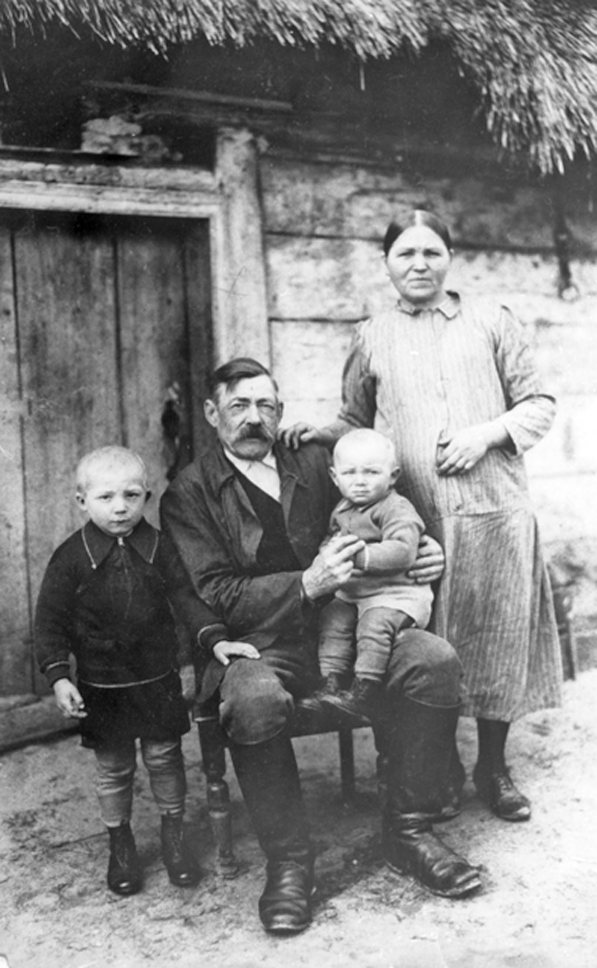 Rok i miejsce wykonania: 1938 r., Naramice, gm. Biała, pow. wieluński, woj. łódzkie.

Fotografia przedstawia postawnego starszego mężczyznę z wąsem siedzącego na krześle, z małym chłopcem na kolanie, przed frontem drewnianej chaty krytej strzechą. Obok niego stoi drugi, kilkuletni chłopiec trzymający rękę na ojcowskim kolanie. Po prawej stronie mężczyzny stoi kobieta, która trzyma rękę na brzuchu i sprawia wrażenie jakby była w zaawansowanej ciąży (?). Zdjęcie ma w sobie dużo uroku, prostoty i siły.

Z informacji nadesłanych przez nadawczynię zdjęcia wynika, że przedstawia ono rodzinę Wawrzyniaków. Od lewej stroi Marian, jej ojciec, a na krześle siedzi jego dziadek Michał, trzymający na kolanach Jana, młodszego brata ojca. Obok stoi babcia Katarzyna. Rodzina pozuje na tle swojego domostwa, które nadawczyni zdjęcia określiła „chatą pradziadków”. Dzieci przebywały u dziadków, ponieważ ich rodzice wyjechali ze wsi za pracą.

Autor zdjęcia: wędrowny fotograf.

Zdjęcie ze zbiorów prywatnych, nadesłała: Leokadia Wawrzyniak z Ząbkowic Śląskich, pow. ząbkowicki, woj. dolnośląskie.

Nr kat. 173-1