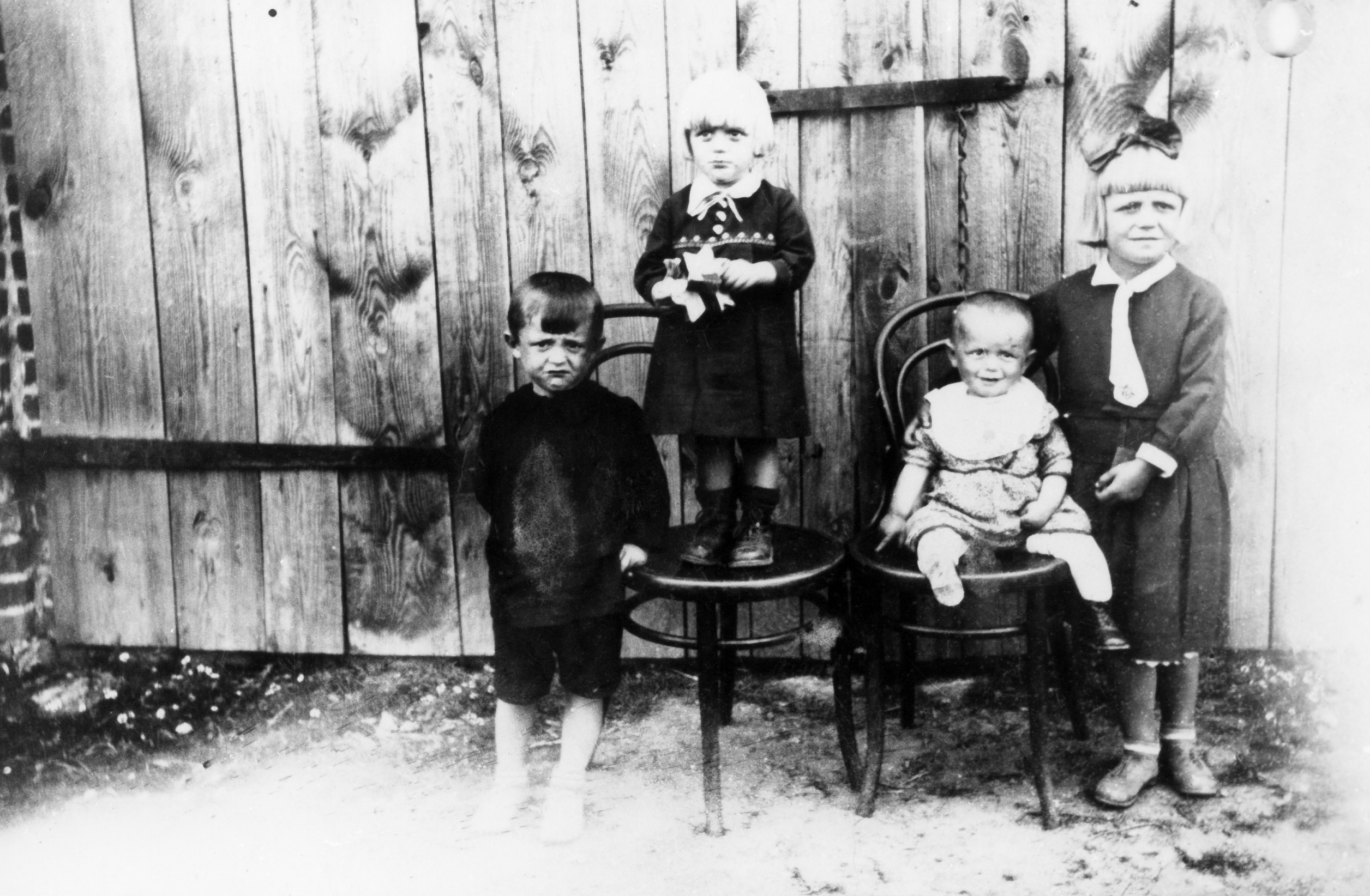 Rok i miejsce wykonania: 1934 r., Racendów, gm. Sławoszew, pow. jarociński, woj. wielkopolskie. Fotografia przedstawia czworo dzieci w różnym wieku. Po lewej stronie stoi mniej-więcej dwuletni chłopiec w ciemnym sweterku i spodniach do kolan. Obok niego, na krześle po prawej stronie, stoi jasnowłosa dziewczynka, ubrana w sukienkę z kołnierzykiem, zaś po jej prawej stronie, też na krześle, siedzi około roczna dziewczynka w jasnej sukieneczce, podtrzymywana przez najstarsze dziecko – dziewczynkę w sukieneczce z białym kołnierzykiem i krawatką, z dużą kokardą we włosach. Zdjęcie zrobiono na tle szerokich drewnianych wrót. Przedstawia ono dzieci teściów nadawczyni, w tym również jej męża. Autor zdjęcia: nieznany. Zdjęcie ze zbiorów prywatnych, nadesłała: Władysława Szymkowiak z Racendowa, k. Sławoszewa, pow. jarociński, woj. wielkopolskie. Nr kat. 277-12