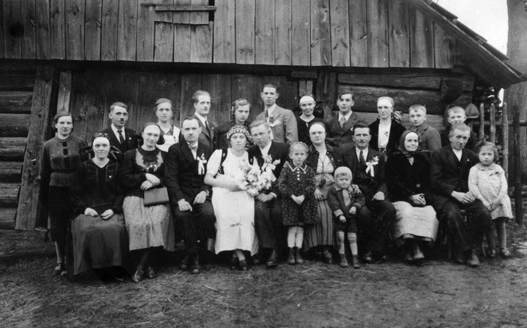 Rok i miejsce wykonania: 1940 r., Łazy na Śląsku Cieszyńskim, gm. Jasienica, pow. bielski, woj. śląskie.

Fotografia przedstawia kilkunastoosobowa grupę osób pozujących na tle dużego drewnianego budynku. W pierwszym rzędzie, pośrodku, siedzą nowożeńcy, obok i za nimi zaproszeni goście, w tym pięcioro dzieci. Panna Młoda ubrana jest w strój regionalny, cieszyński, widoczny jedynie fragmentarycznie: ma kabotek, czyli białą koszulę z bufiastymi rękawami, ciemniejszy żywotek (rodzaj gorsetu) i białą zapaskę (fartuch). Włosy ma upięte (słabo widoczne), a na nich wianek. W rękach trzyma bukiet kwiatów. Pan Młody nachyla głowę w jej stronę. Ubrany jest w ciemny garnitur, w którego klapę wpięty ma mały bukiet przybrany kokardą. Podobne bukiety pozostali mężczyźni. Wśród pozujących do zdjęcia jest sporo dzieci. Większość kobiet nosi stroje regionalne, a na głowach ma charakterystyczne dla Śląska Cieszyńskiego czepce.

Według właściciela zdjęcia przedstawia ono siostrę matki, Annę Sławińską i Franciszka Kocmana w dniu ich ślubu. Zdjęcie zostało wykonane na tle zabudowań gospodarczych rodziny Sławińskich we wsi Łazy.

Autor zdjęcia: fotograf z Zakładu Fotograficznego „REKORD” w Bielsku.

Zdjęcie ze zbiorów prywatnych rodzin Sławińskich i Kocmanów.

Nr kat. 214-1