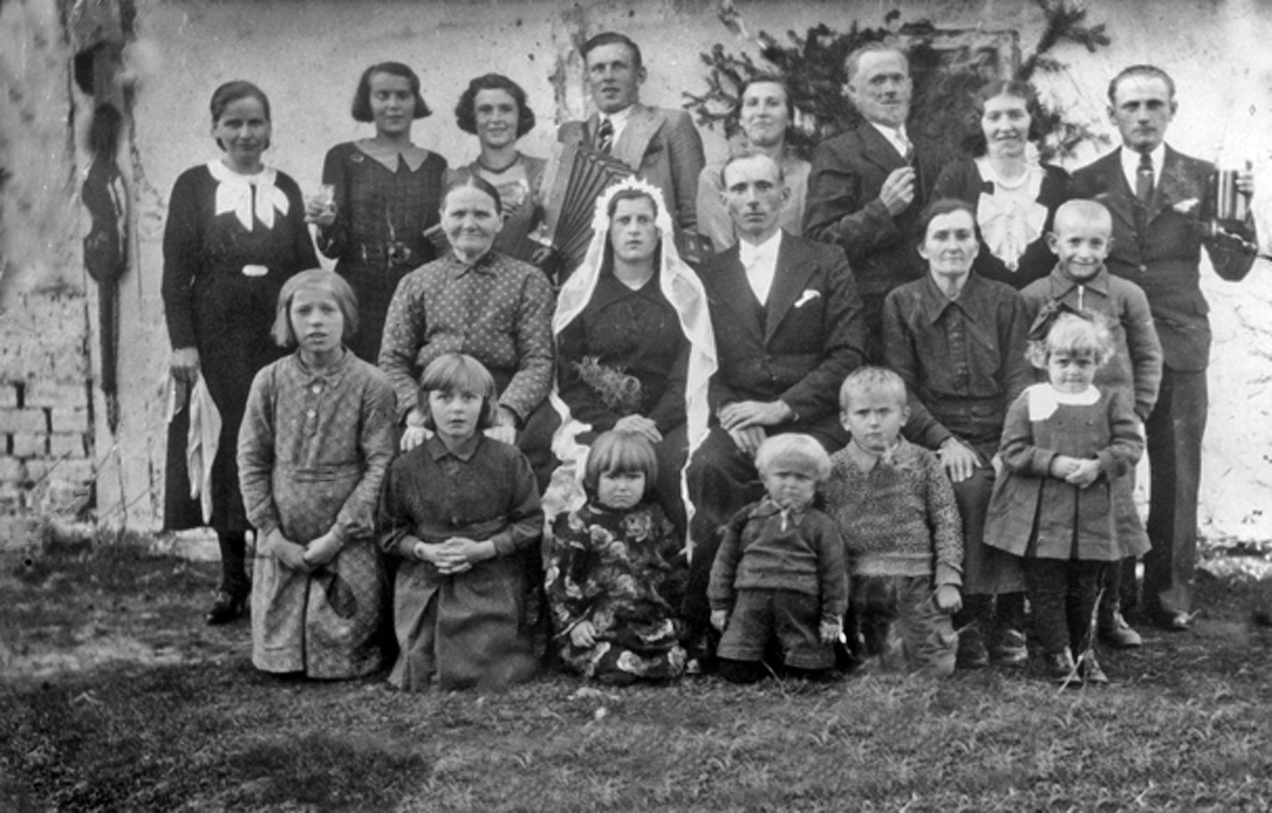 Rok i miejsce wykonania: sierpień 1937 r., Domatowo (kaszb. Domôtowò), gm. Puck, pow. pucki, woj. pomorskie.

Fotografia przedstawia grupę dziewiętnastu osób, w tym siedmiorga dzieci w różnym wieku, pozujących do zdjęcia na tle drewnianego, pobielonego budynku z wejściem udekorowanym gałęziami świerkowymi. Goście weselni pozują do zdjęcia w trzech rzędach, przy czym pierwszy rząd składa się z pięciorga klęczących dzieci i jednej stojącej dziewczynki. W drugim rzędzie, pośrodku, siedzi Młoda Para, ubrana w ciemną, odświętna odzież. Kobieta ma na włosach długi biały welon, a mężczyzna nosi w ciemny garnitur i białą koszulę. W trzecim rzędzie stoi osiem osób. Mężczyzna w środku ma przewieszony przez ramię akordeon. Mężczyzna z prawej strony kadru w swojej lewej dłoni trzyma butelkę (jakby z zamiarem napełnienia kieliszka, który trzyma mężczyzna po jego prawej?). Pomiędzy nimi stoi młoda kobieta. Według właściciela zdjęcia przedstawia ono wesele jego wuja.

Autor zdjęcia: nieznany.

Zdjęcie ze zbiorów prywatnych, nadesłał: Jan Okonek, wieś Koślinek-Dąbrówka Malborska, pow. sztumski, woj. pomorskie. 

Nr kat. 242-2